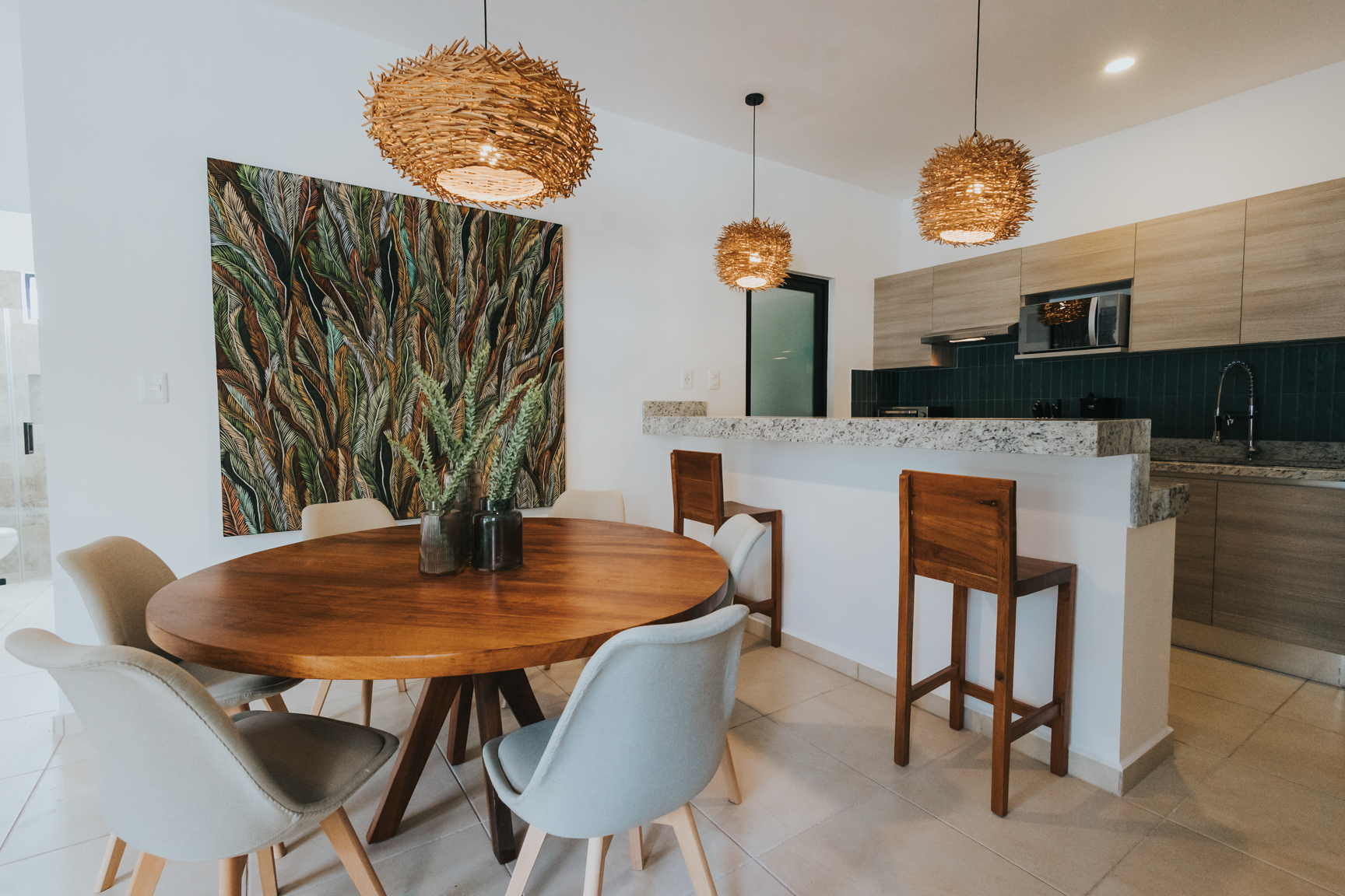 Open dining and kitchen with round wood table, woven pendants and tropical leaf artwork
